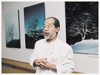 写真家、宮崎学氏による春夏秋冬12枚同じ位置から撮影した柿の樹の写真を背にして説明する鎌田さん